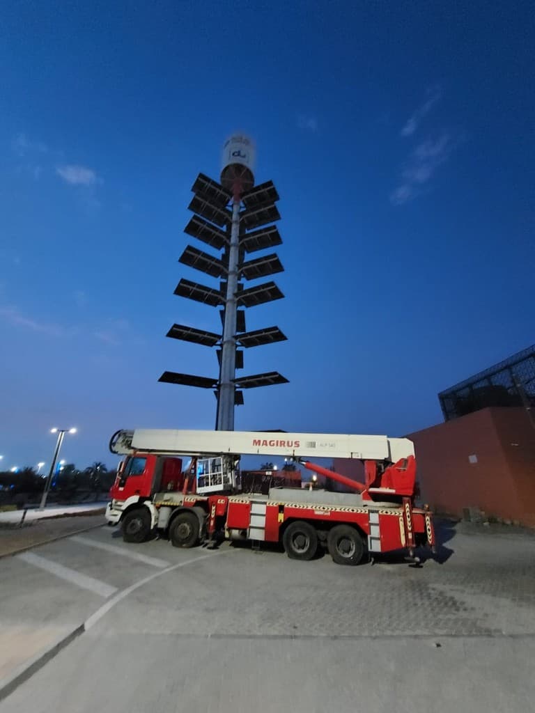 Iveco Magirus truck-mounted manlift beside a lighting tower at dusk